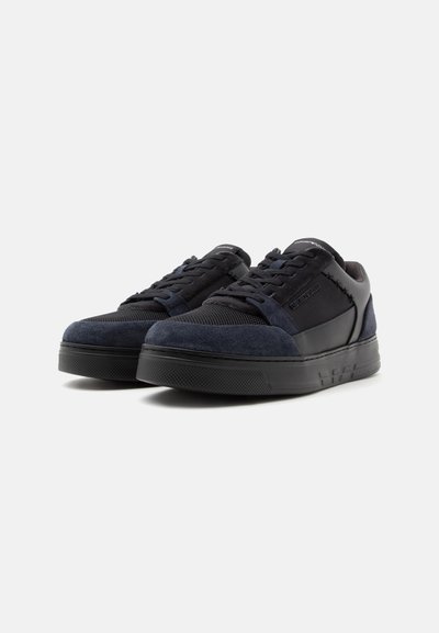Ζευγάρι μαύρα και σκούρα μπλε sneakers χαμηλού τύπου με κορδόνια μπροστά και υφασμένα καουτσούκ πέλματα σε μονόχρωμο ανοιχτό φόντο.