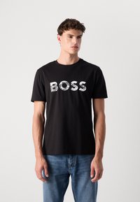 Schwarzes Baumwoll-T-Shirt mit dem Wort "BOSS" in Weiß, wobei einige Buchstaben mit einem Gittermuster versehen sind. Einfacher Rundhalsausschnitt und kurze Ärmel.