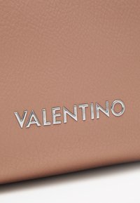 Valentino logo uz rozā teksturētas ādas, ar sudraba metāla burtiem ar gludu virsmu un skaidru redzamību.