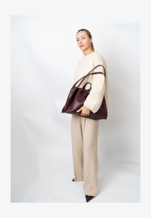 Burgundy leather tote bag with a gathered top, two long handles, and a smooth texture, paired with a cream oversized sweater and beige trousers.