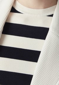 Pull rayé noir et blanc avec texture côtelée, présentant de larges rayures horizontales. Partiellement recouvert par un blazer texturé de couleur crème.