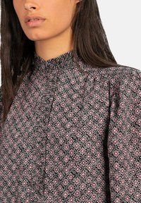 Blusa negra con un alto cuello fruncido y un patrón floral en rosa y blanco, que presenta acentos plisados y una tela texturizada.