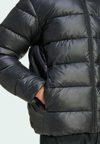 Läheltä kuvattu musta untuvatakki, joka esittelee sen tikattua pintarakennetta ja osittain vetoketjulla kiinni olevaa etuosaa, käsi lepuuttaa lantiolla.