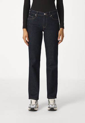 Person iført mørkeblå jeans med lige ben, sort langærmet top og hvide-grå sneakers, stående mod en ensartet lys baggrund.