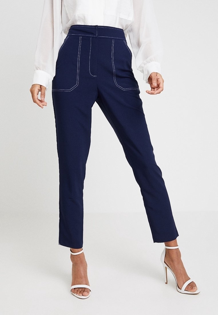 Woman wearing navy tailored pants with white contrast stitching, sheer white blouse, and white high-heeled sandals standing on white floor.