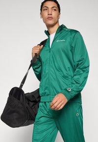 Πράσινη αθλητική φόρμα από γυαλιστερό ύφασμα, με ζακέτα με φερμουάρ και στενά παντελόνια με ελαστικές μανσέτες και λεπτομέρειες με λογότυπο. Μαύρη τσάντα duffel.