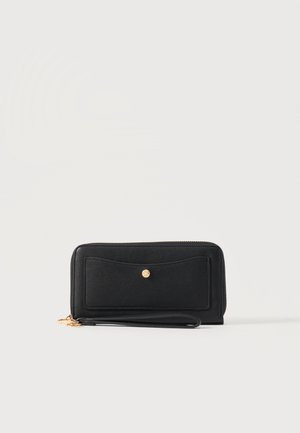 Portefeuille bracelet noir zippé avec poche avant et petit bouton doré, posé sur fond blanc.