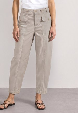 Pantalon cargo beige avec poches à rabat boutonnées, porté avec un haut blanc et des sandales plates noires avec des accents dorés sur un sol gris.
