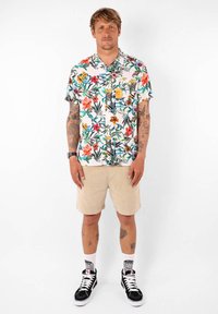Chemise à manches courtes à motifs floraux avec un fond clair ; shorts en kaki ; baskets montantes noires ; chaussettes blanches avec logo.