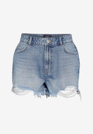 Femme debout portant une chemise rouge vif à manches courtes, un short en jean bleu usé et des baskets marron, posant avec une main dans la poche.