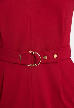 Zbliżenie czerwonego paska materiałowego z podwójną złotą klamrą i dwoma złotymi ćwiekami na pasującym czerwonym ubraniu.