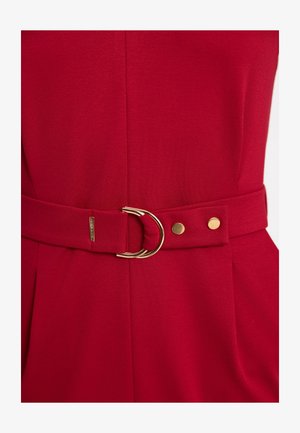 Zbliżenie czerwonego paska materiałowego z podwójną złotą klamrą i dwoma złotymi ćwiekami na pasującym czerwonym ubraniu.