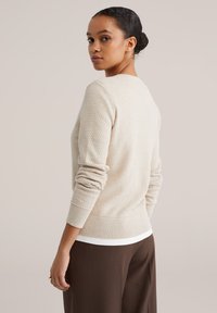 Svetloružový žíhaný sveter s dlhými rukávmi, rebrovanými manžetami a lemom. Nosený cez biely top, doplnený o hnedé nohavice. Hladká tkanina.