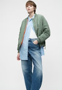 Asmuo, vilkintis žaliu bomber stiliumi švarku virš mėlynos dryžuotos marškinių ir baltos bluzės, derinamas su laisvais mėlynais džinsais, stovi, su viena ranka kišenėje.