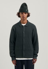 Jonge man in een donkergroene, gestructureerde, knoop cardigan, donkergroene gebreide muts, witte onderbroek en donkere broek, staande tegen een effen achtergrond.