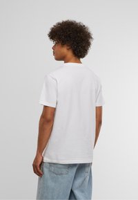 Een persoon met krullend haar, gekleed in een eenvoudig wit T-shirt en lichtblauwe spijkerbroek, staat met de rug naar de camera tegen een grijze achtergrond.
