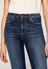 Mörkblå jeans med slim fit, featuring hög midja, framfickor och subtila sömnadsdetaljer. Slät textur, denimmaterial.