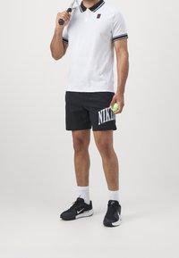 Mies tennis-pelaaja valkoisessa poolopaidassa, mustissa Nike-shorteissa ja mustissa tennareissa, pitää tennismailaa olkansa yli ja palloa kädessään.