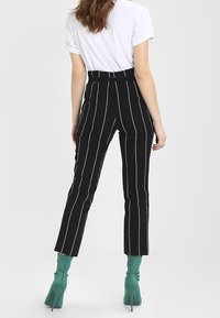 Černobílé pruhované kalhoty s vysokým pasem a zúženými nohavicemi, spárované se zelenými kotníkovými botami, na jednobarevném pozadí.