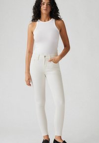 Jeans ajustados de mezclilla blanca con cintura alta, que cuentan con cierre de botón y bolsillos delanteros, combinados con una camiseta sin mangas blanca ajustada.