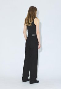 Jeans anchos negros de mezclilla, con una cintura alta, dos bolsillos traseros y una etiqueta en la cintura. Estilizados con una camiseta sin mangas negra.