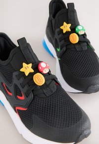Chaussures de sport noires avec semelles blanches, présentant des touches rouges et vertes et décorées de breloques à thème Super Mario sur les lacets.