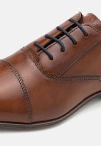 Chaussure en cuir marron avec une finition lisse, bout arrondi, détails de couture subtils et lacets noirs passés à travers six œillets.
