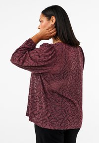 Maglione lungo a maniche lunghe color marrone scuro, con una texture e un motivo di forme astratte. Presenta una vestibilità comoda e un orlo rotondo, abbinato a pantaloni neri.
