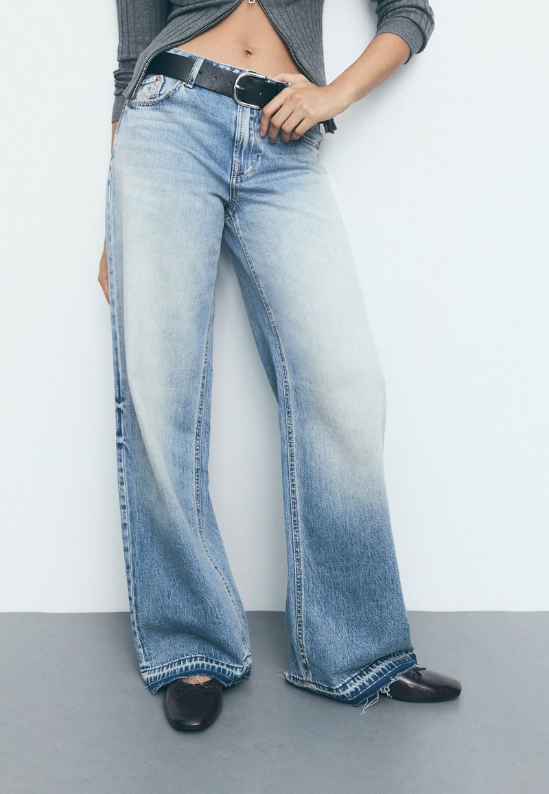 Personne portant un jean large bicolore avec des ourlets effilochés, une ceinture noire, des chaussures foncées et un haut court gris zippé.