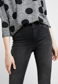 Suéter gris con grandes lunares negros, combinado con jeans negros de tiro alto. La tela parece suave con un acabado ligeramente texturizado.