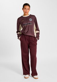 Bordeauxrotes Langarmshirt mit vertikalen Streifen und Logo, kombiniert mit passenden, locker sitzenden bordeauxroten Hosen. Das Shirt verfügt über cremefarbene Akzente und Bündchen.