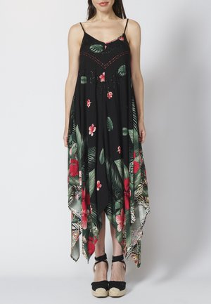 Kobieta ubrana w czarną, bezrękawnikową sukienkę z tropikalnym czerwono-zielonym kwiatowym wzorem i asymetrycznym dołem, w połączeniu z czarnymi sandałami na koturnie espadryl.