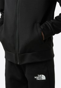 Čierna bunda na zips s dvomi bočnými vreckami, vyrobená z hladkej látky; na ľavej strane zodpovedajúcich nohavíc je biely logo.