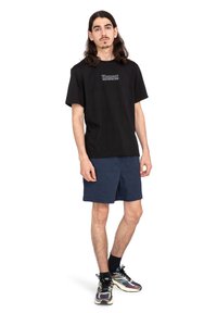 Svart bomullströja med tryckt logo, i kombination med marinblå shorts. Modellen har mönstrade träningsskor och står i en neutral position.