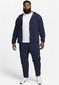 Marine atletiekjack met een ritssluiting aan de voorkant, capuchon en zijzakken, gecombineerd met bijpassende taps toelopende broeken en witte sneakers. Glad stoftextuur.