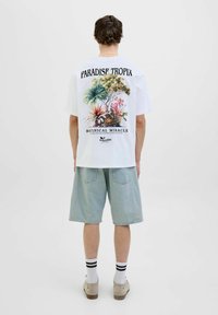 Hvid kortærmet T-shirt med et botanisk print på ryggen, teksten "PARADISE TROPIX" og "BOTANICAL MIRACLE", parret med lyseblå denimshorts.