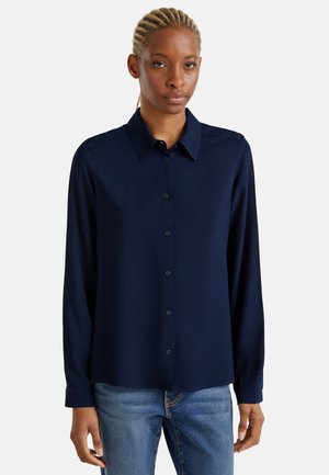 Frau mit geflochtenem Haar, die ein marineblaues, langärmeliges Hemd mit Knopfleiste und blaue Jeans vor einem schlichten hellen Hintergrund trägt.