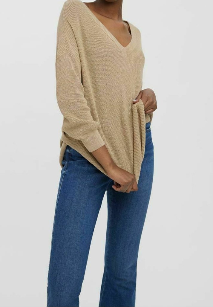 Personne portant un pull beige ample à col en V et un jean bleu, tenant le bord inférieur du pull avec les deux mains.