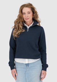 Felpa con zip blu navy, maniche lunghe, texture morbida e collo alto, indossata sopra una camicia bianca. Design casual con vestibilità morbida.