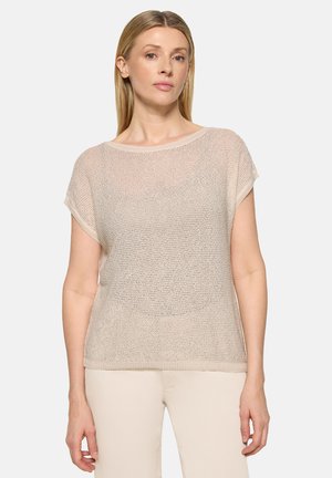 Kvinde med glat blondt hår iført en lysebeige strikbluse med korte ærmer og cremefarvede bukser, stående foran en ensfarvet baggrund.