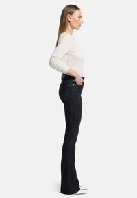 Jean flare noir avec poches avant et couture discrète, associé à un pull ajusté de couleur claire, vu de côté.