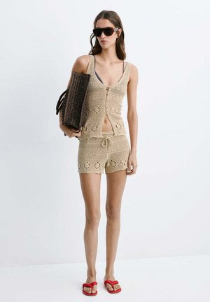Mujer con top de crochet beige y pantalones cortos, sosteniendo un bolso tejido oscuro, con gafas de sol grandes y chanclas rojas, de pie sobre un fondo blanco liso.