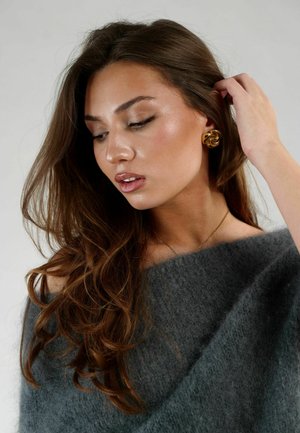 Maglione grigio, morbido e texture, scivola da una spalla. Riccioli provocatori incorniciano il viso, ornato da orecchini d'oro a spirale. Pelle liscia, trucco neutro.