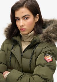 Olive green puffer jacket with fur-lined hood, black and white drawstrings, and a red Superdry Everest patch on the sleeve. Textured stitching visible.