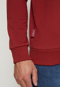 Bordeauxfarbenes Sweatshirt mit gerippten Bündchen und Saum, ausgestattet mit einem Seitenlabel. Weiche Textur, schlanker Schnitt und kontrastierende Nähte.
