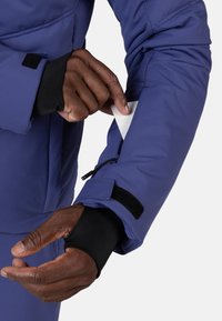 Fioletowa ocieplana kurtka z zapinaną na zamek kieszenią na ramieniu, czarne polarowe mankiety widoczne. Ręka wkładająca białą kartę do kieszeni.