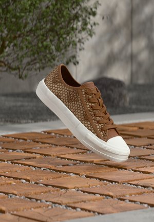 Bruine sneaker met zeshoekig gaaspatroon, witte rubberen zool en neuskap, zwevend boven houten en kiezelstenen buitenvloer.