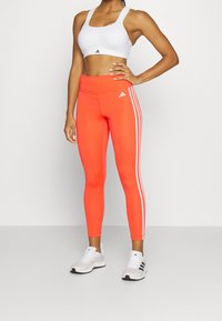 Leggings de sport rouges avec trois bandes verticales blanches sur les côtés, associés à un soutien-gorge de sport blanc et des baskets blanches. Texture de tissu lisse.