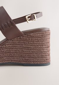 Sandale compensée marron avec plateforme en corde tressée, bride en cuir à la cheville et détail de boucle dorée sur fond neutre.