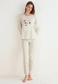 Lichtbeige pyjama-set met een longsleeve top met een hondenapplicatie en een broek met een patroon van kleine zwarte hartjes.